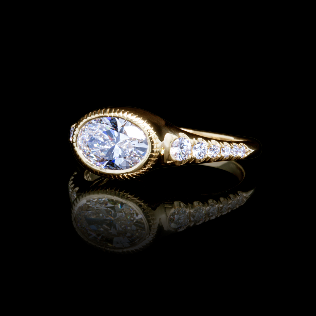Handcrafted oval diamond engagement ring in 18K yellow gold with pavé detailing, artistic reflection shot.