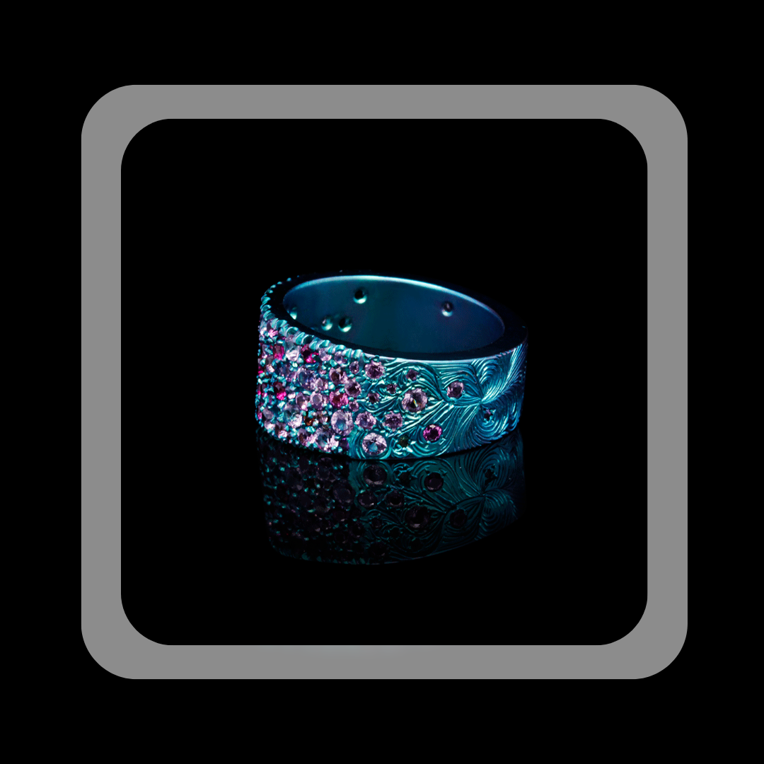 A close-up of a titanium ring featuring pavé-set la pink sapphires and engraved feather-like patterns, displayed against a reflective black background. The design highlights the craftsmanship this high-jewelry piece.
