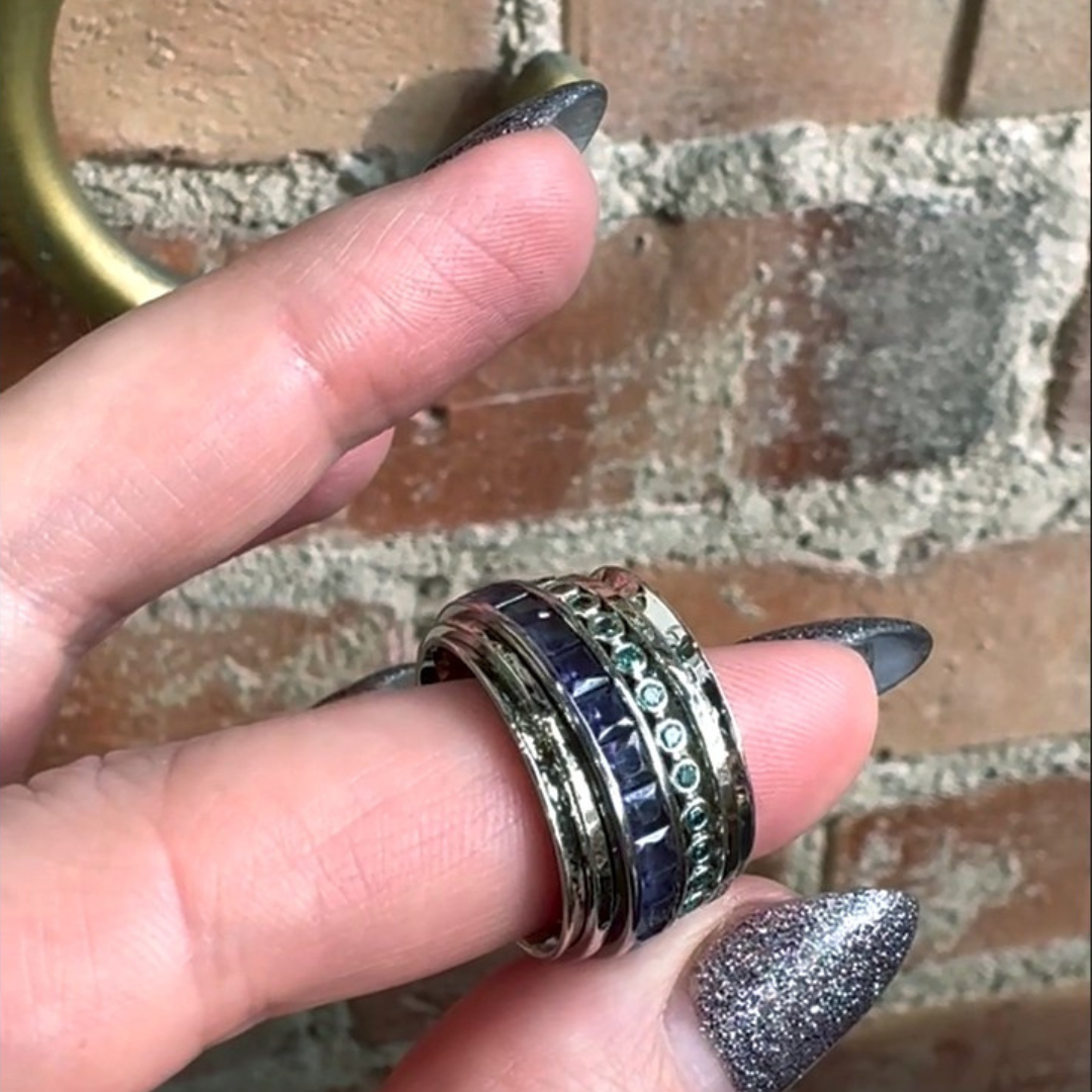hand holding fidget ring and getting ready to fiddle with it. featuring 4 spinning bands on wide base ring. round blue diamonds and purple sapphire princess cuts included in the design