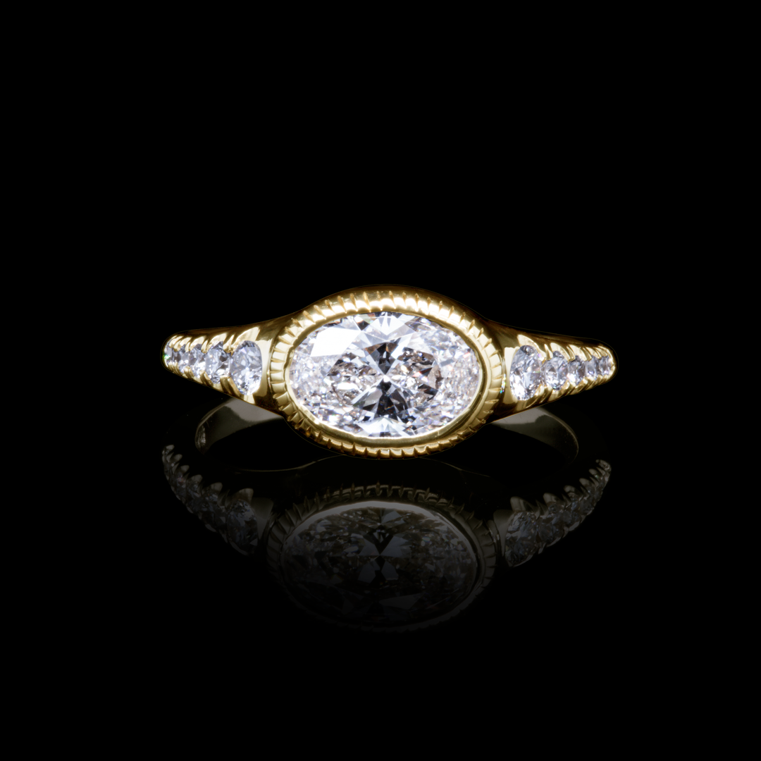Oval diamond engagement ring in yellow gold with side stones, photographed on black background for contrast.