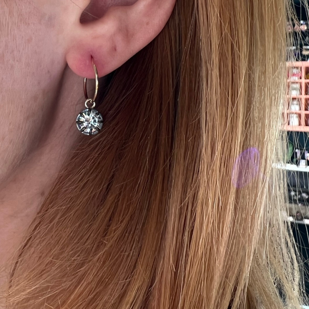 A close-up side view of an ear wearing a gold hoop earring with a dangling solitaire gemstone set in a silver prong setting. The gemstone catches the light, showcasing its brilliance and sparkle.