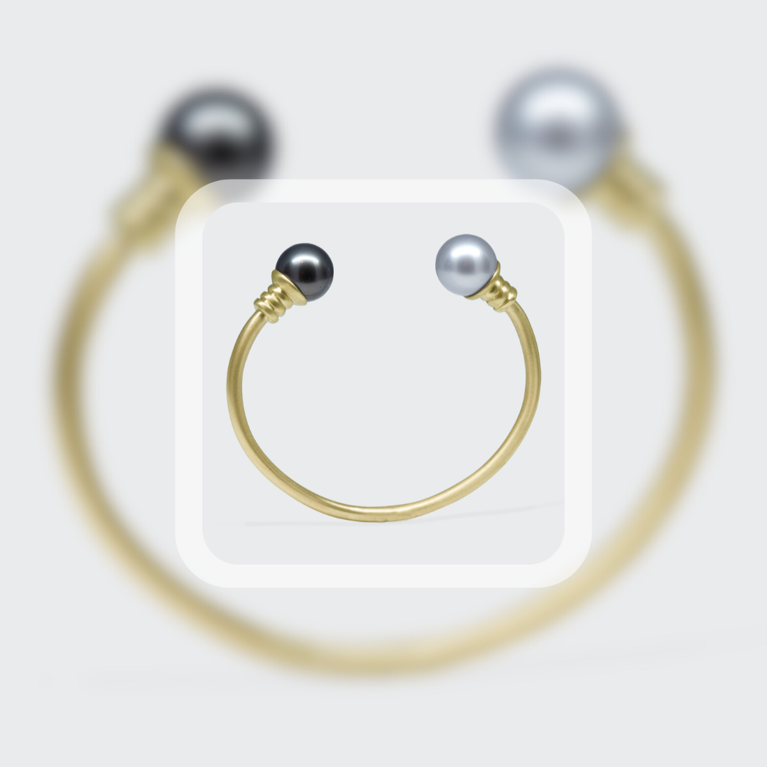 A close-up of a gold cuff bracelet with black and white pearls on its ends, showcased against a minimalist grey background with blurred reflections for emphasis on the jewellery’s elegance.