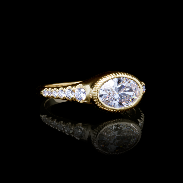 Yellow gold oval diamond ring with pavé diamonds, side angle on reflective black background.