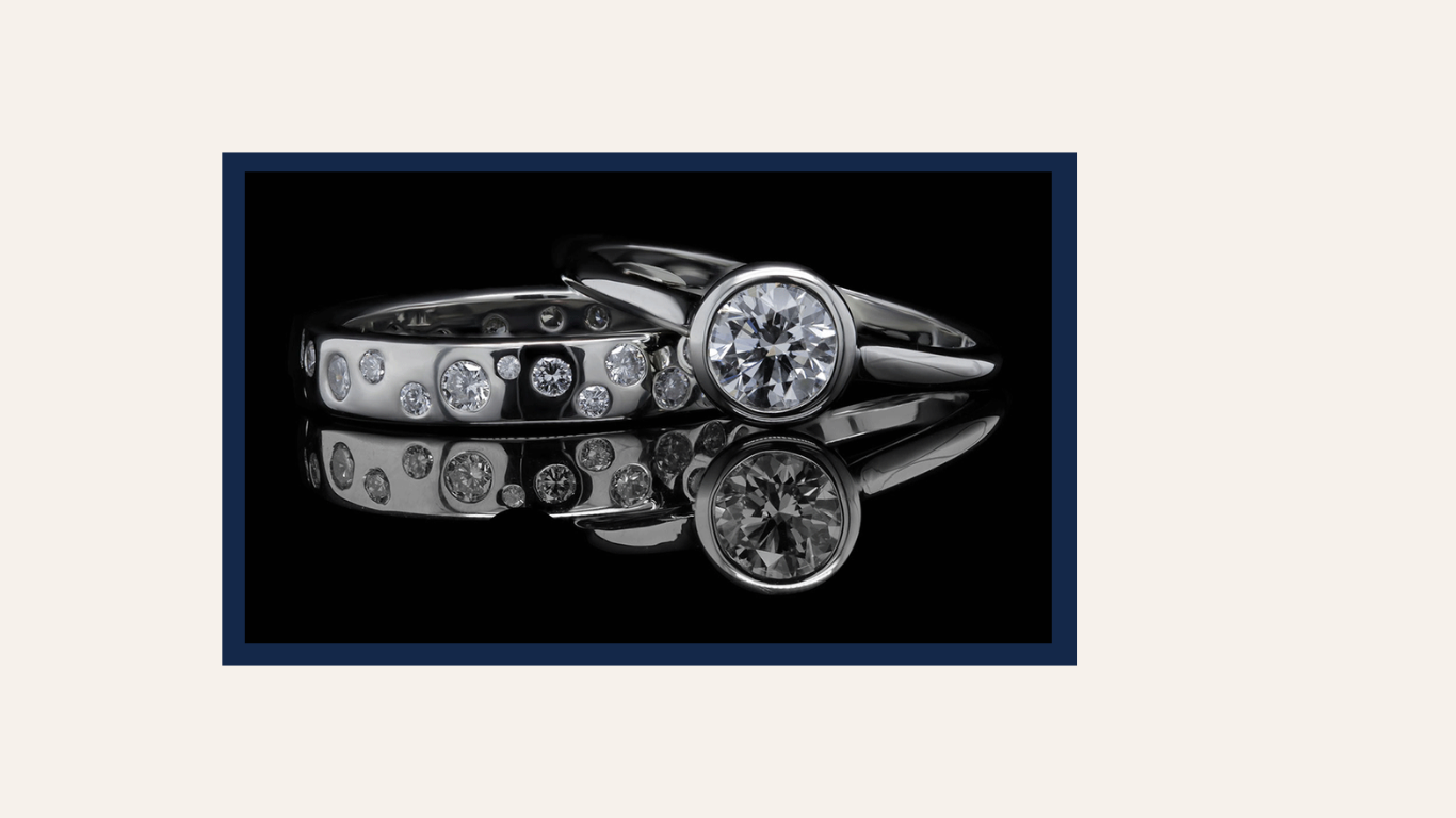 A stunning pair of platinum rings, one featuring a bezel-set round diamond with sleek, modern lines, and the other adorned with scattered flush-set diamonds. The rings are displayed against a reflective black background, emphasizing their brilliance and elegant craftsmanship.