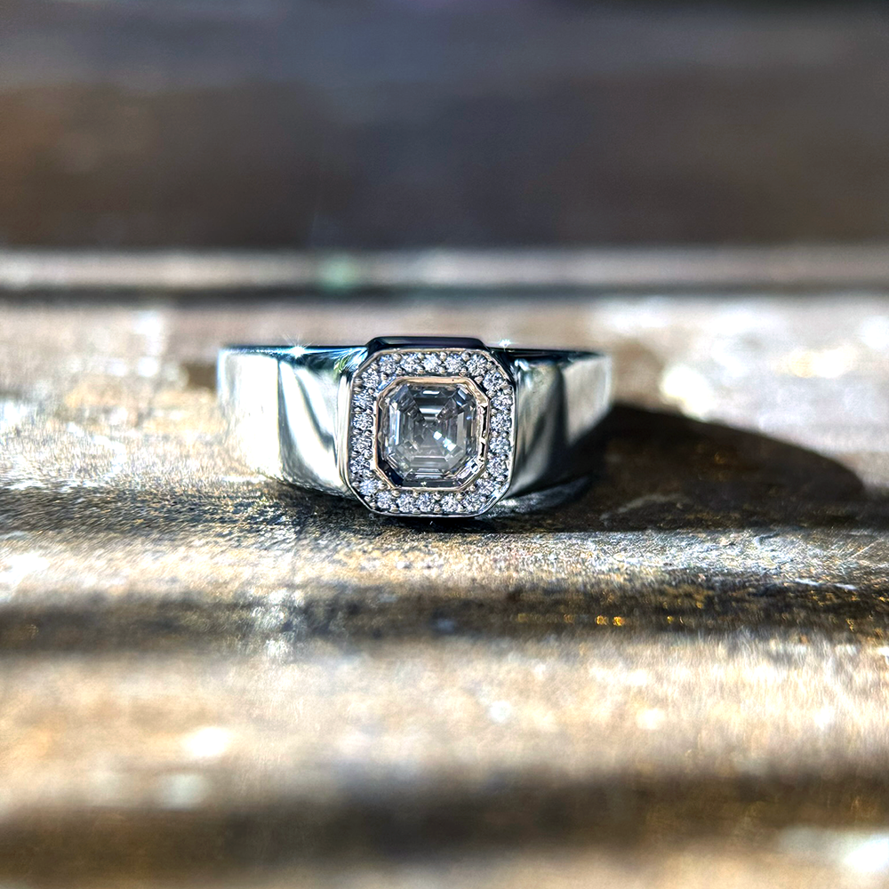 18 karat white gold ring sitting on a sunlit background. Ring features an asscher-cut center stone, surrounded by a natural diamond halo. The shank is wide and high-polished with sparkling diamonds