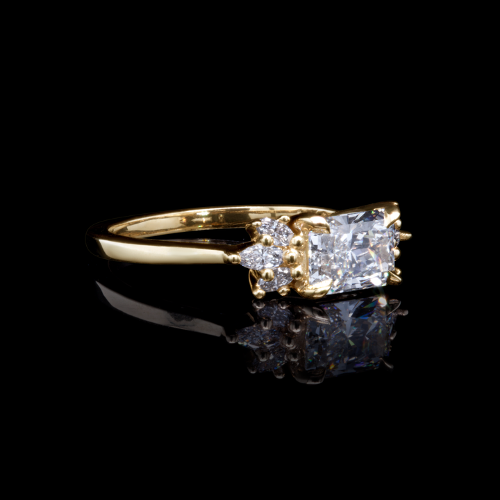 Radiant cut lab diamond engagement ring with marquise accents in 14K yellow gold, artistic black background.