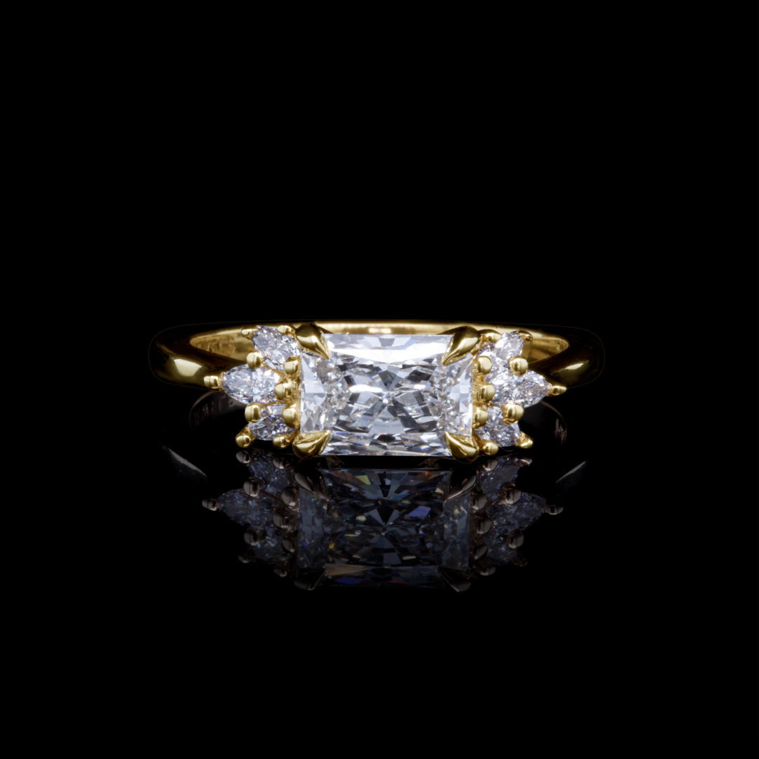 Radiant cut diamond engagement ring in yellow gold with side stones, photographed on black background for dramatic contrast.