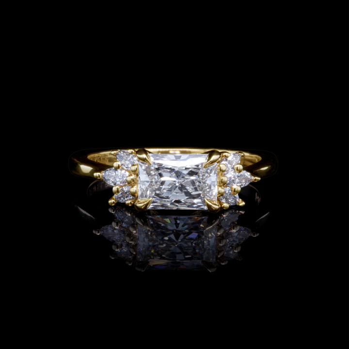 Radiant cut diamond engagement ring in yellow gold with side stones, photographed on black background for dramatic contrast.