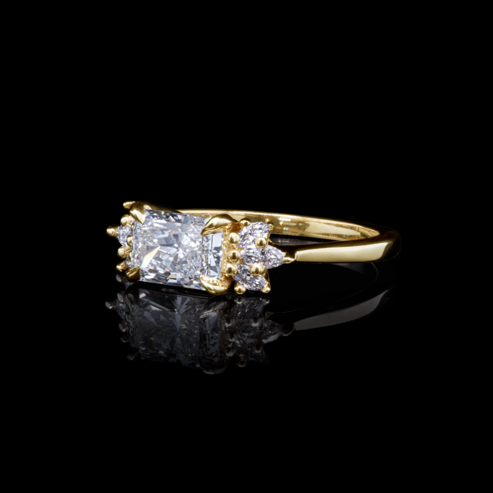 Handcrafted radiant diamond engagement ring in recycled yellow gold with marquise and round side stones, reflection image.