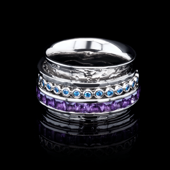 A detailed product shot of a white gold stackable ring set featuring blue gemstones in a bezel setting and square-cut purple stones displayed against a reflective black background. The rings showcase a mix of textures and vibrant colours.