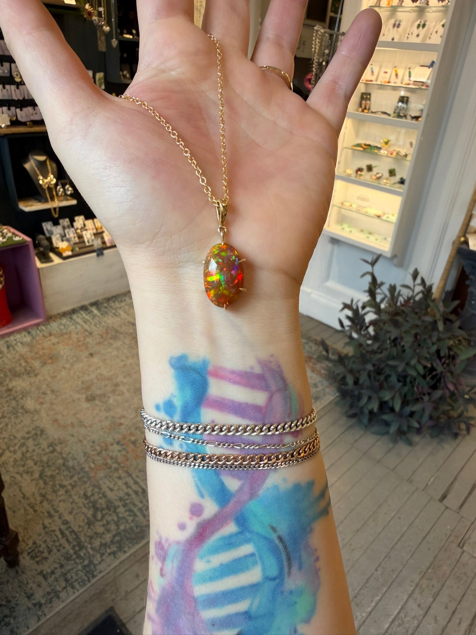 Hand wearing a gold necklace chain with an opal pendant, with tattoos visible on the wrist, in an indoor setting.