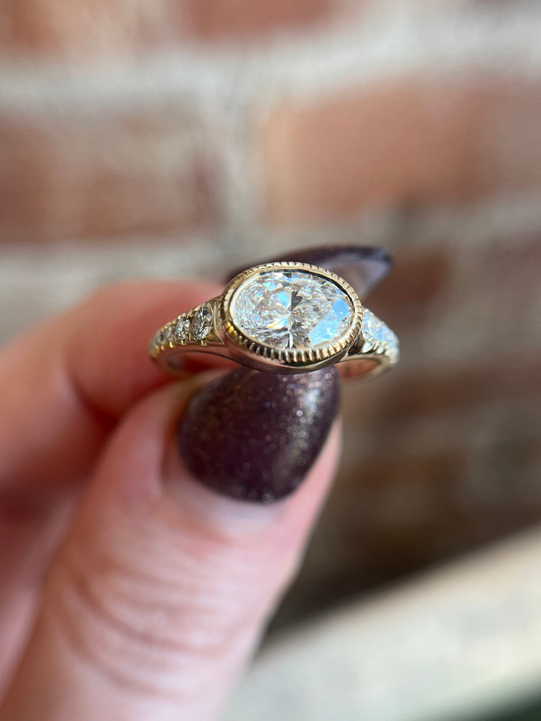 Oval diamond engagement ring in yellow gold with diamond accents, worn on model’s hand in natural lighting.