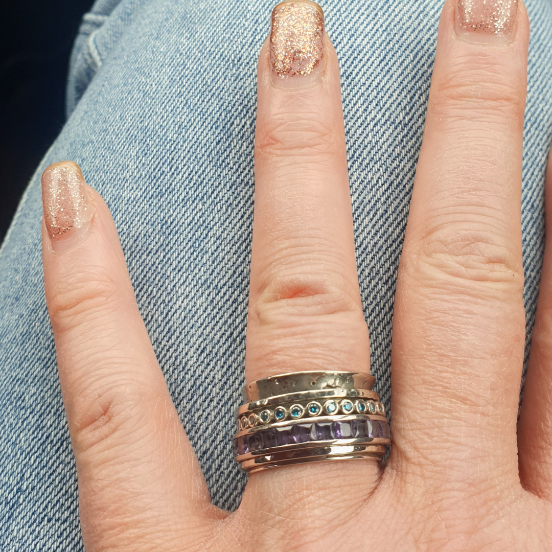 A close-up of a hand wearing a stack of white gold rings with intricate details, including blue gemstones and purple stones, set against a casual denim background. The nails are polished with a glittery gold finish.