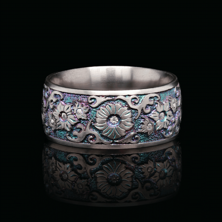 Close-up of iridescent titanium ring with hand-engraved floral design and bezel-set white moissanite stones, displayed on a black background.