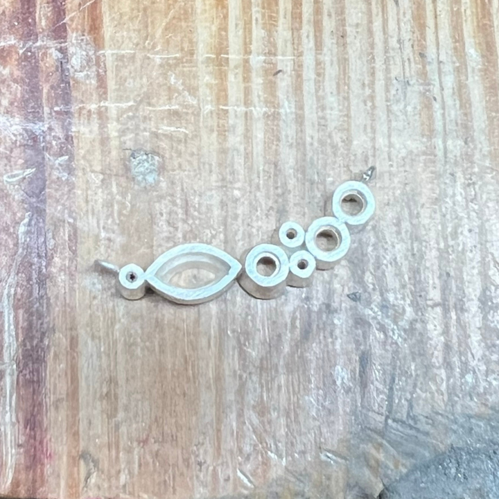 An unfinished necklace in the making, crafted in silver, showing the open bezels and framework for setting marquise-cut and round gemstones, laid on a wooden jewellery bench.