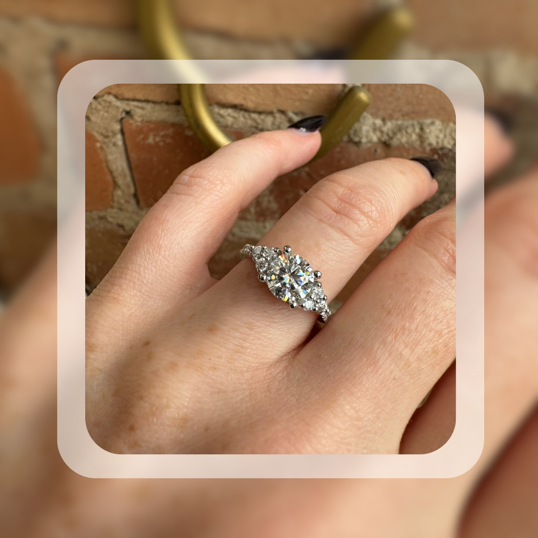 A platinum engagement ring with an round moissanite center stone, flanked by shimmering moissanite accents on a delicate band. 