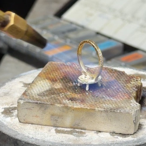A gold ring is being soldered with a torch in the background.