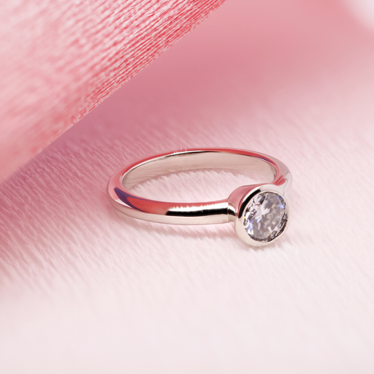 Close-up of a classic solitaire ring. The band is made of 18K white gold and features a single, sparkling moissanite gemstone in the center. Ring is sitting on top of a pink background.